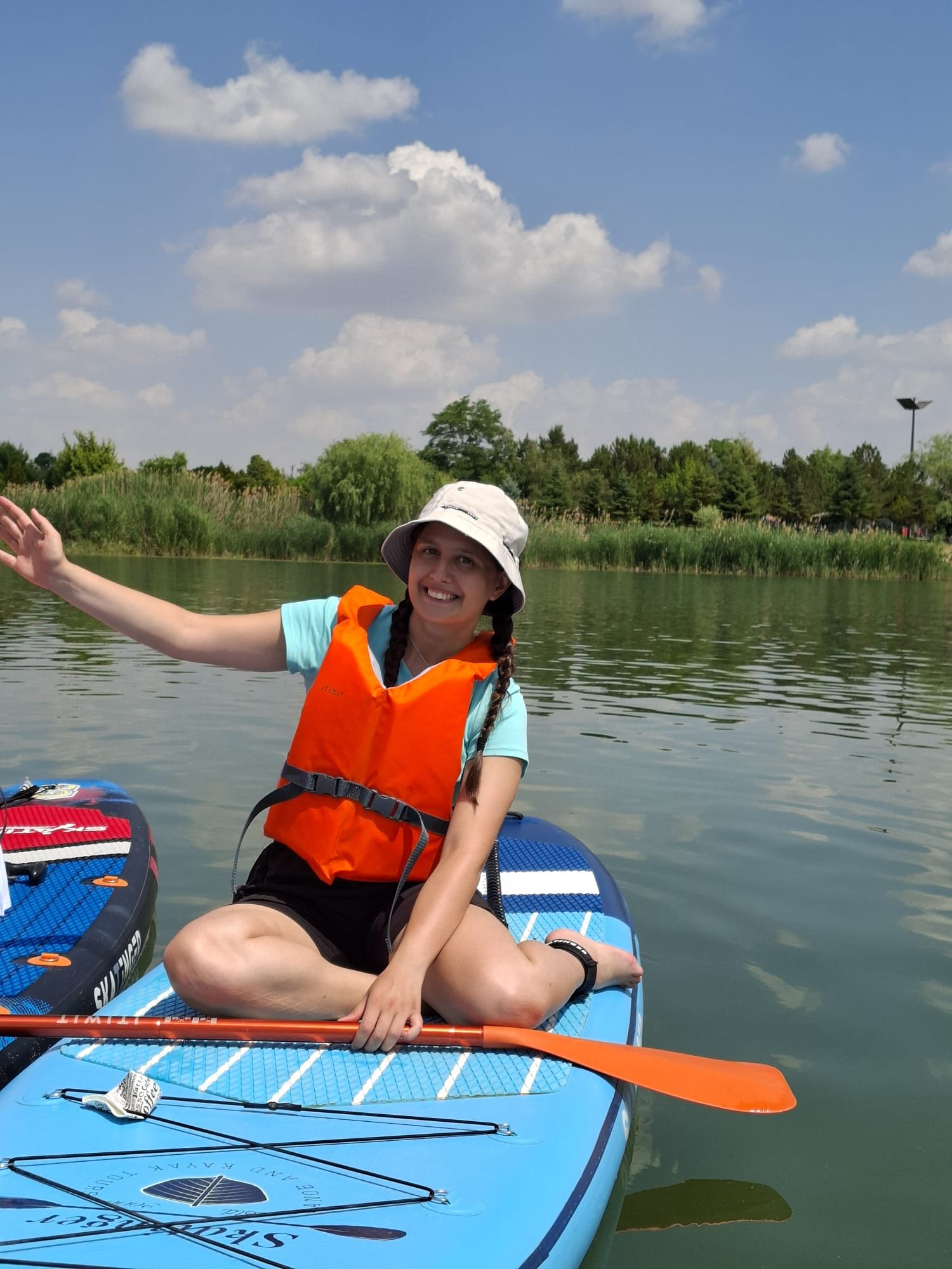 About Deniz sitting on a blue stand-up paddleboard on calm water, wearing an orange personal flotation device and a sun hat, holding a paddle