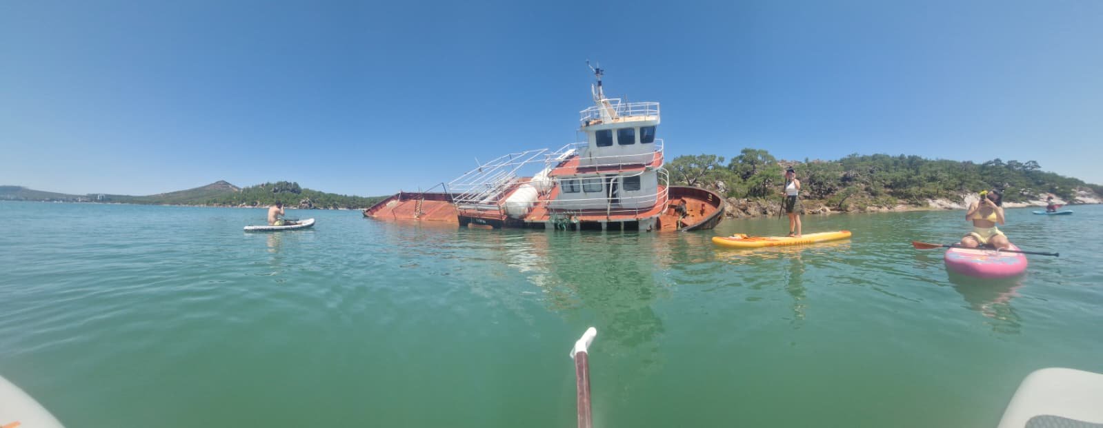 Paddleboarding in Ayvalık: The Shipwreck Route to Delikli Taş Group of paddleboarders on inflatable SUP boards gathered around a half-sunken shipwreck in clear turquoise water at Ayvalık, Turkey