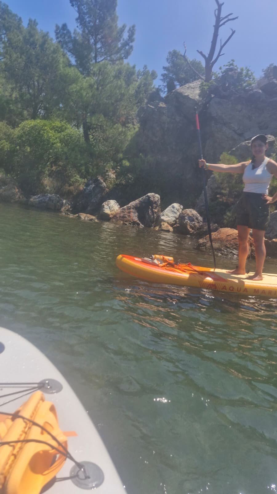 Paddleboarding in Ayvalık: The Shipwreck Route to Delikli Taş Paddler standing upright on an orange Aqua Marina inflatable paddleboard in clear calm water beside rocks and pine trees on the Ayvalık coast