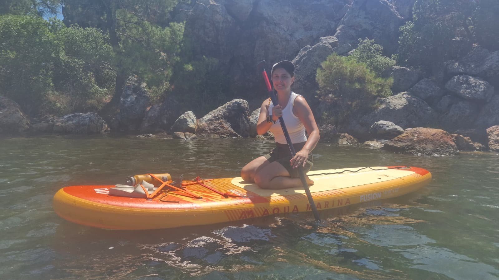 Paddleboarding in Ayvalık: The Shipwreck Route to Delikli Taş Paddler kneeling on an orange Aqua Marina Fusion inflatable paddleboard with water bottle and gear strapped to the deck, beside rocks on the Ayvalık coast