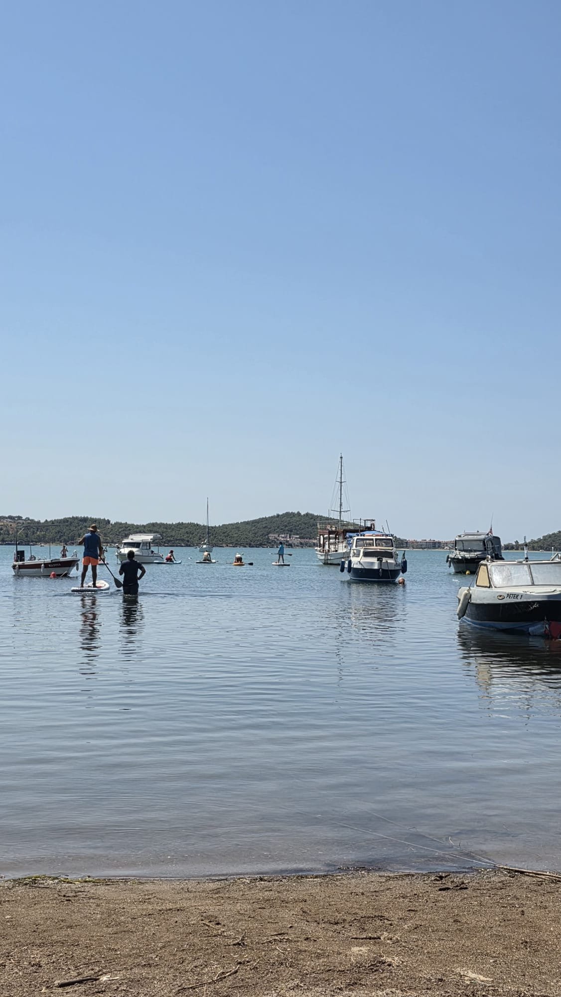Paddleboarding in Ayvalık: The Shipwreck Route to Delikli Taş Paddleboarders setting off from the sandy beach at Çamlık, Ayvalık, weaving between moored boats on completely flat water