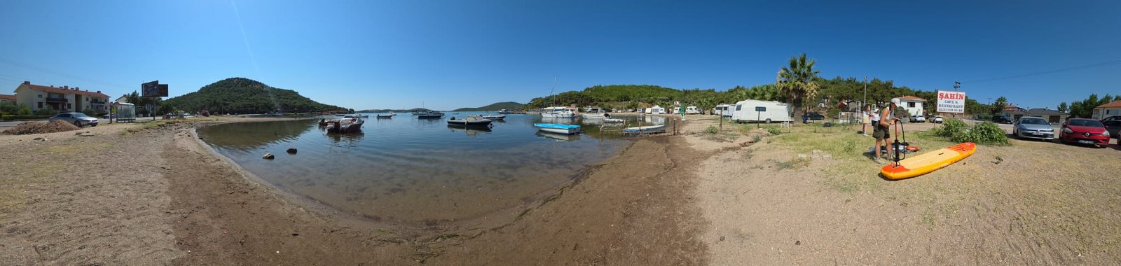 Paddleboarding in Ayvalık: The Shipwreck Route to Delikli Taş Panorama of the paddleboard launch beach at Çamlık, Ayvalık, with an inflatable SUP being pumped up on the sand, moored fishing boats and pine-covered hills behind