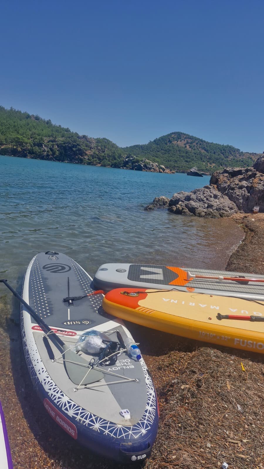 Paddleboarding in Ayvalık: The Shipwreck Route to Delikli Taş Three inflatable paddleboards pulled up on a rocky shore at Ayvalık with paddles, leashes and water bottles secured to the decks