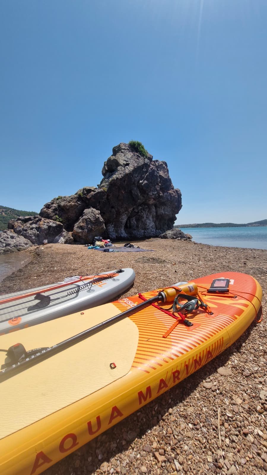 Paddleboarding in Ayvalık: The Shipwreck Route to Delikli Taş Delikli Taş, a dark volcanic rock with a natural hole through its base, rising above a pebble beach at Ayvalık, with inflatable paddleboards and a snorkel mask in the foreground