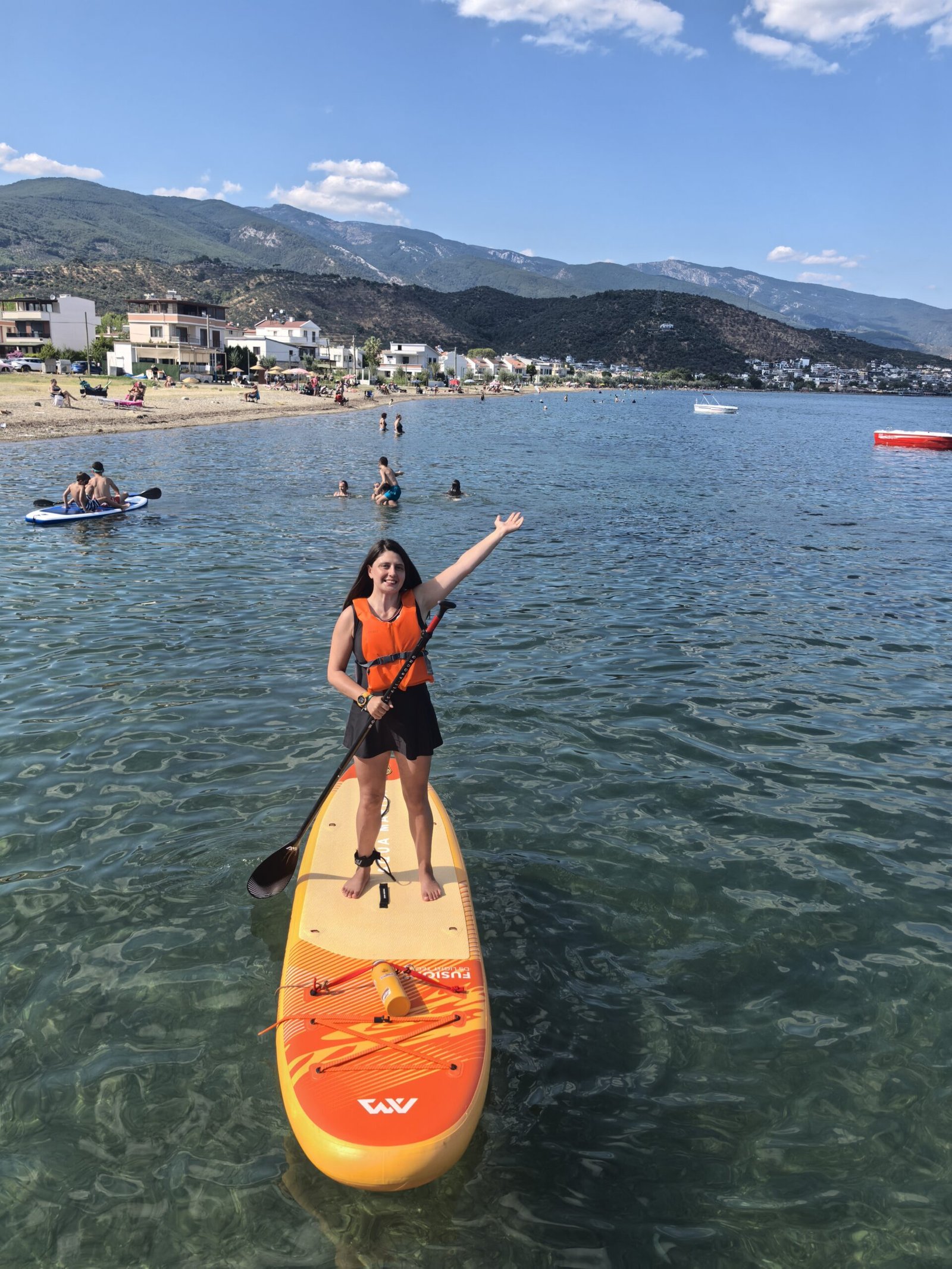 Deniz Ulaşan standing on an orange stand-up paddleboard in clear shallow water, wearing an orange PFD and an ankle leash, with the coastline and mountains behind